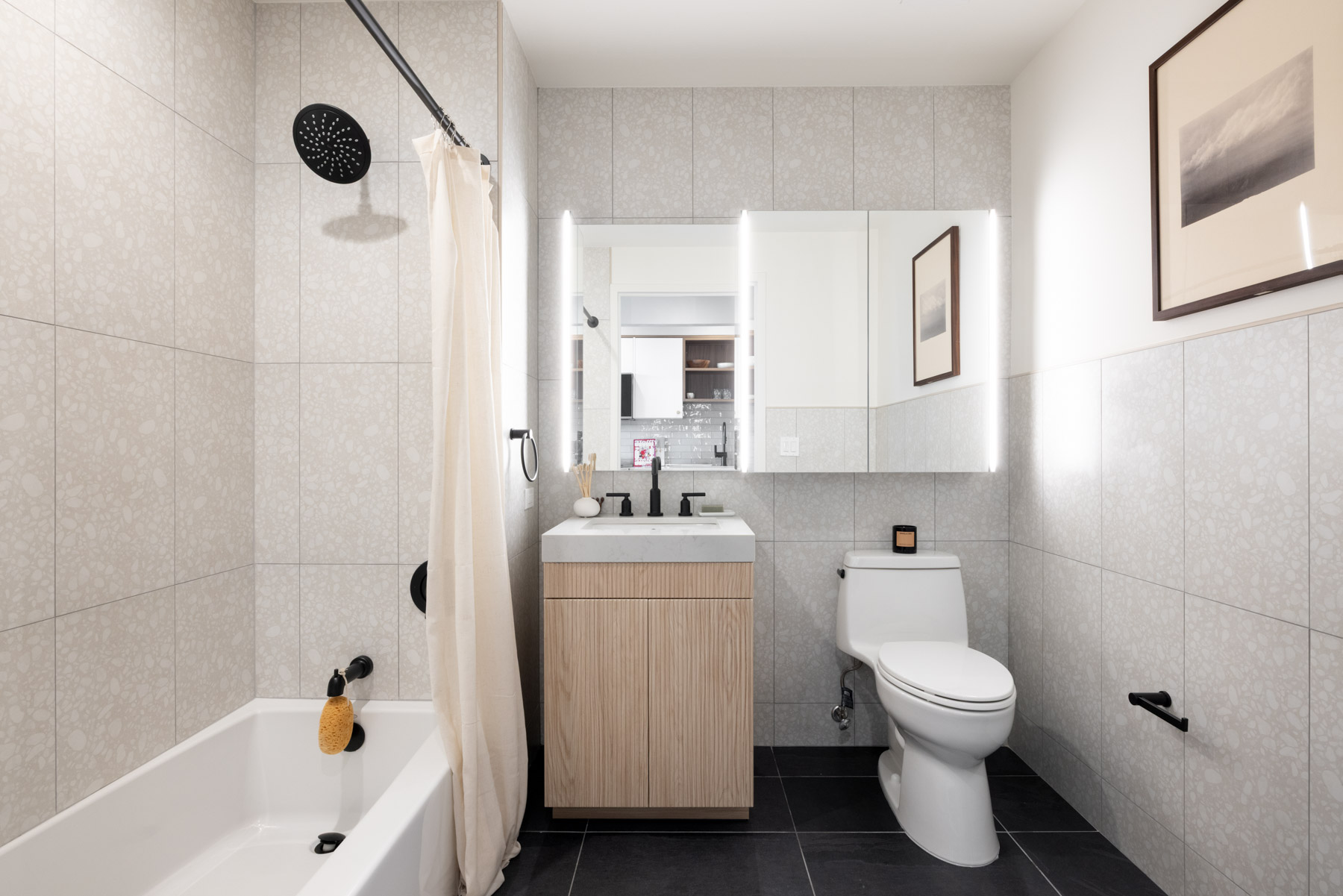 Modern bathroom with bath/tub combo, Light Wood Laminate vanity with London Grey Stone Countertops, white tub with dark fixtures, and Medicine Cabinets with Accent Lighting