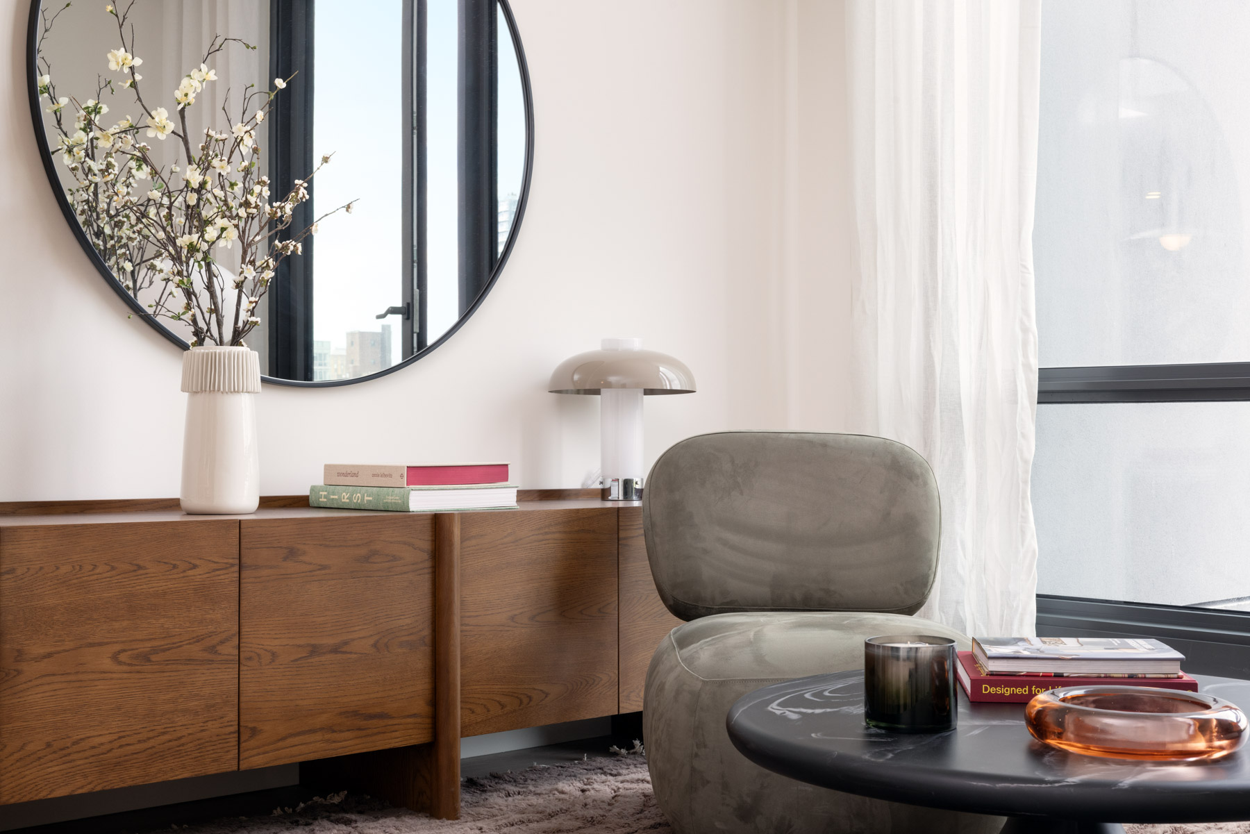 Detail of media cabinets with flowers in vase, books and lighting, large round mirror above and coffee table with candle and books in front