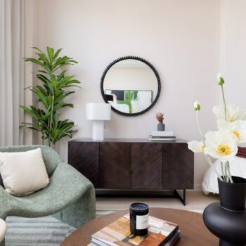 Detail of coffee table, vase with flowers, seating and media cabinet with floor plant to the side and round mirror on wall