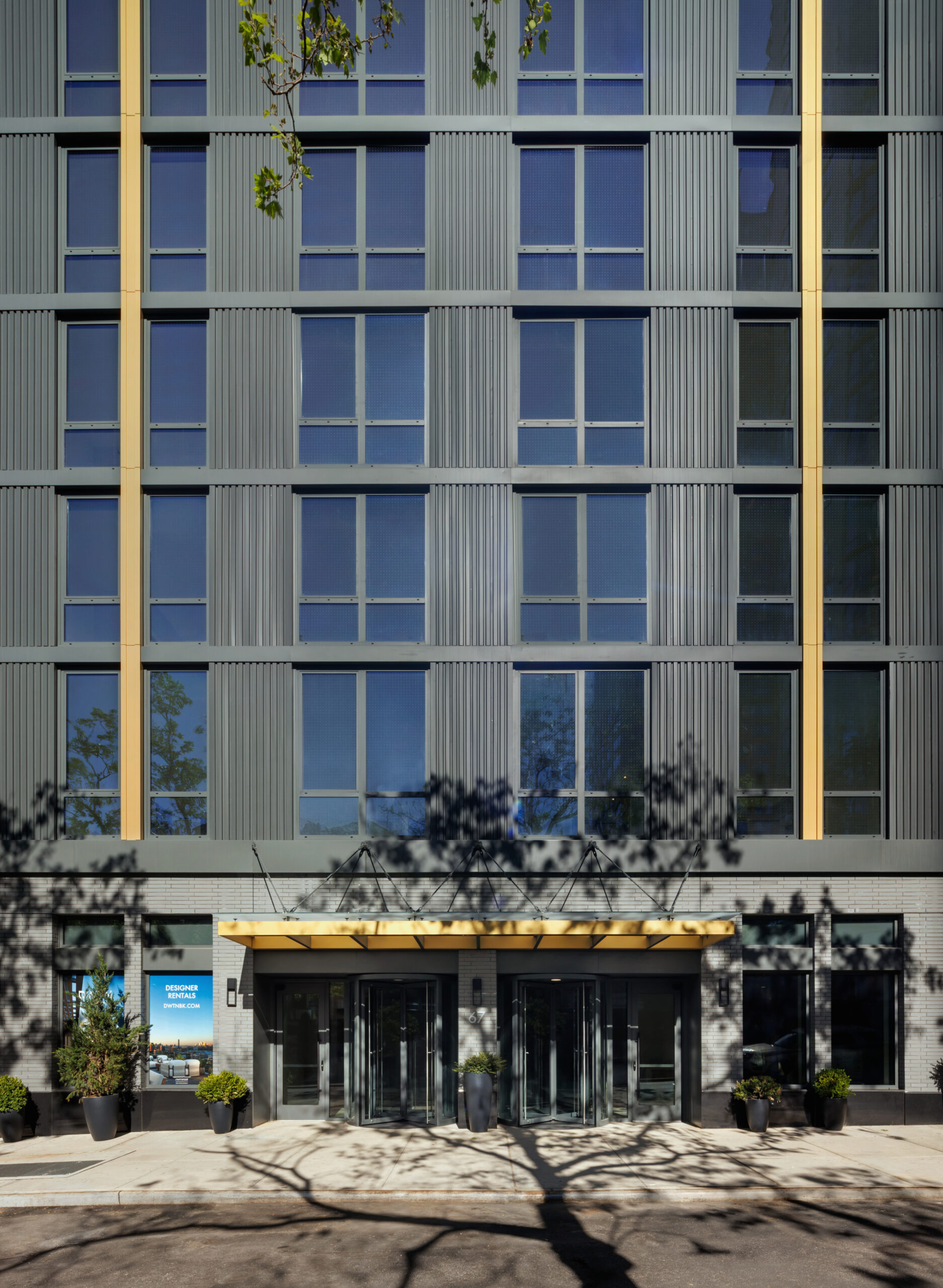Street-level view of a modern apartment building entrance featuring a gray corrugated facade with vertical gold accents, double revolving glass doors under a wide gold canopy, and potted greenery flanking the sidewalk.