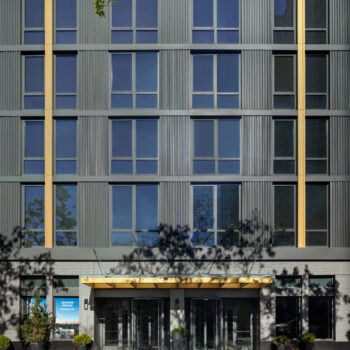 Street-level view of a modern apartment building entrance featuring a gray corrugated facade with vertical gold accents, double revolving glass doors under a wide gold canopy, and potted greenery flanking the sidewalk.