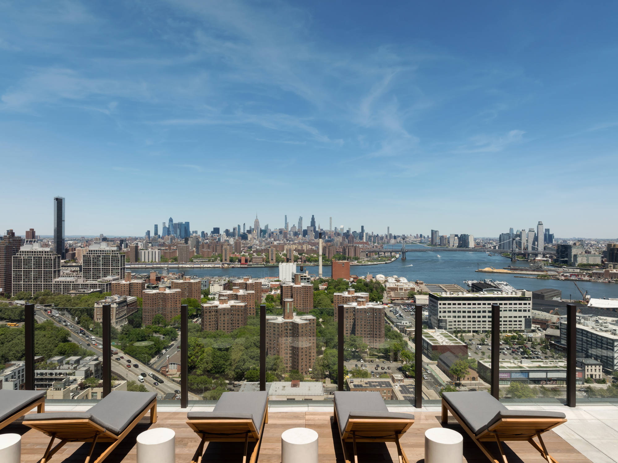 Rows of sleek wooden lounge chairs lined up on a rooftop terrace behind a glass safety railing, offering an expansive, wide-angle view of the city river, distant bridges, and the urban skyline.