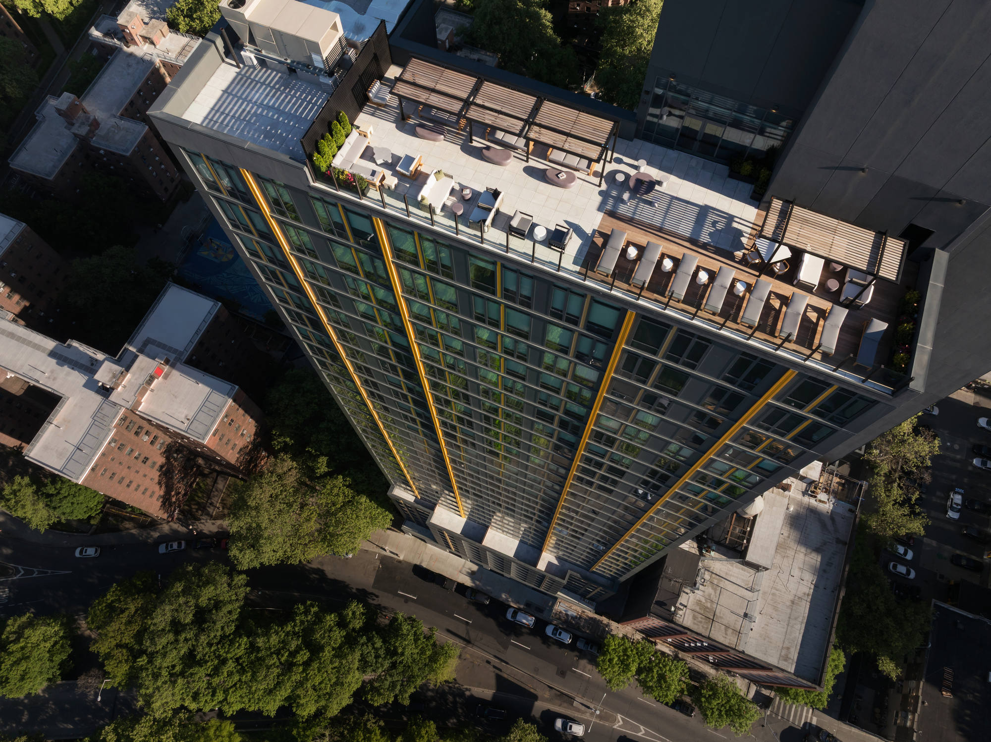 Birds-eye aerial view looking straight down at the luxury apartment building's fully furnished rooftop deck, showcasing organized lounge seating zones, pergolas, and the surrounding city streets below.