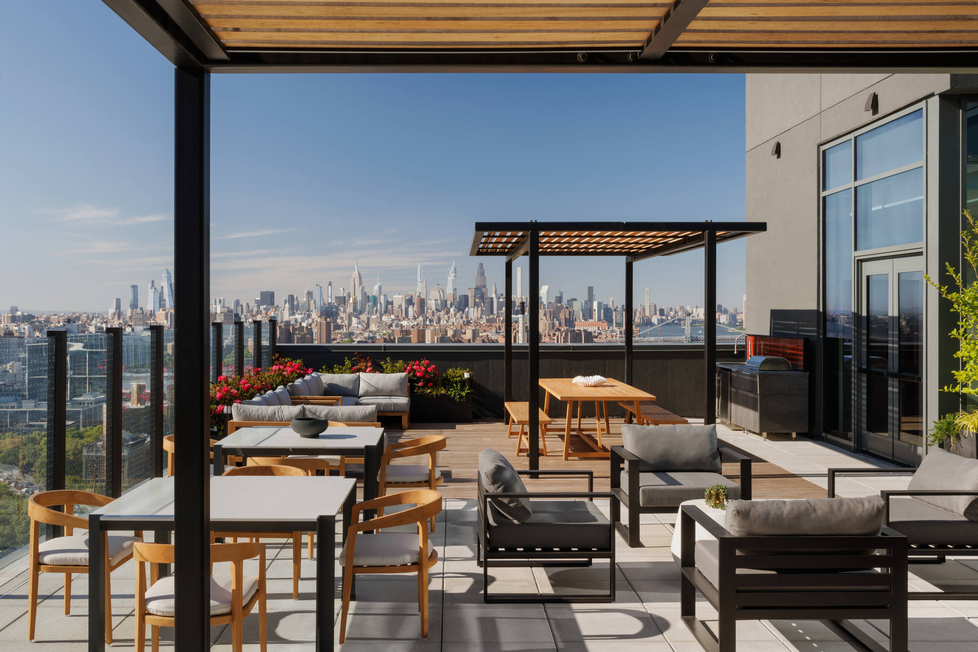 Rooftop dining and entertainment area featuring high-end outdoor tables, cushioned armchairs, a covered wooden pergola, and an outdoor kitchen station against a backdrop of the distant city skyline.