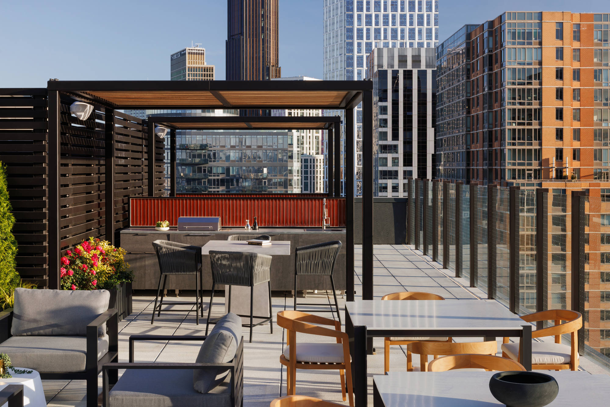 Close-up of a premium rooftop amenity space featuring a modern outdoor grill station with a red tile backsplash, high-top bar seating, and dining tables enclosed by a glass railing.