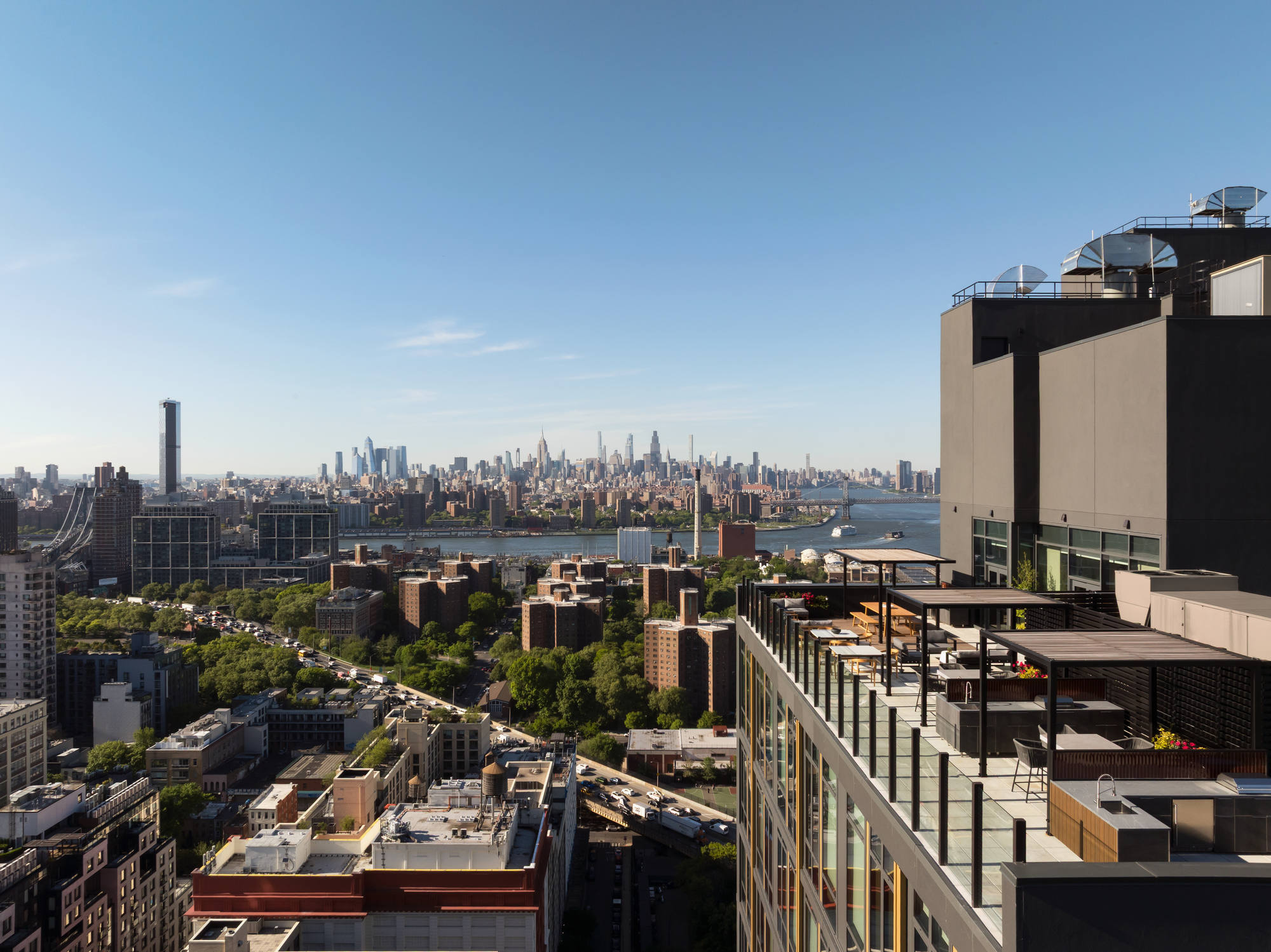 High-angle scenic shot looking past a luxury building's multi-level rooftop terrace down toward a bustling urban landscape, tree-filled parks, and a distant city skyline under a clear sky.