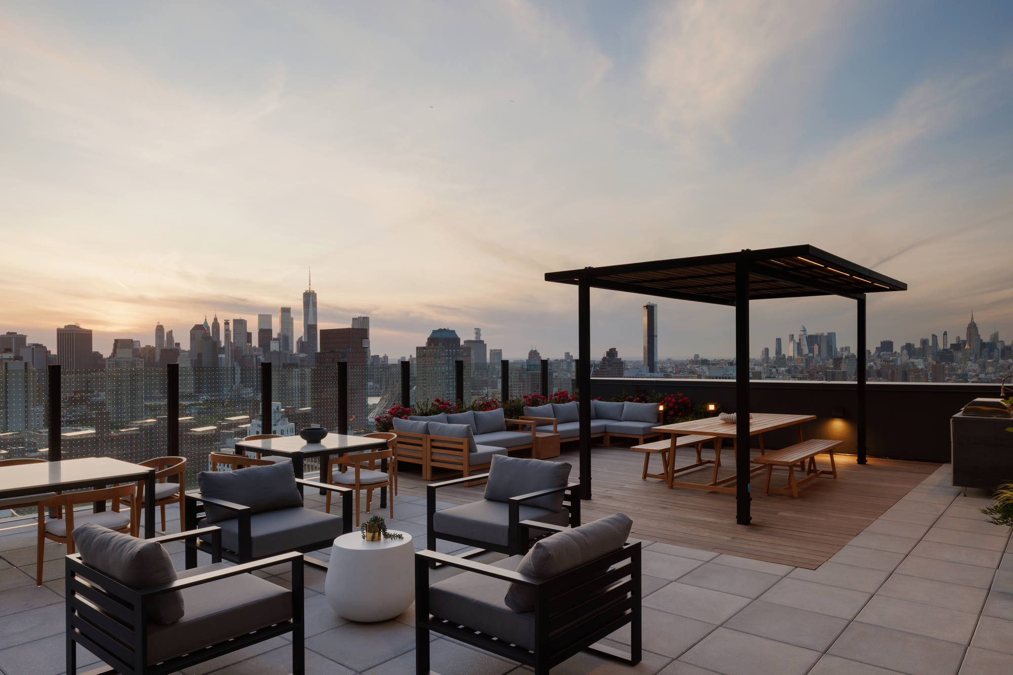Luxury rooftop lounge at sunset featuring modern gray armchairs, a long wooden picnic table under a black metal pergola, and a sleek glass railing framing a view of the distant city skyline.