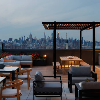 Modern rooftop amenity deck at twilight showcasing upscale patio furniture, an outdoor kitchen station, and subtle under-bench accent lighting beneath a wooden pergola.