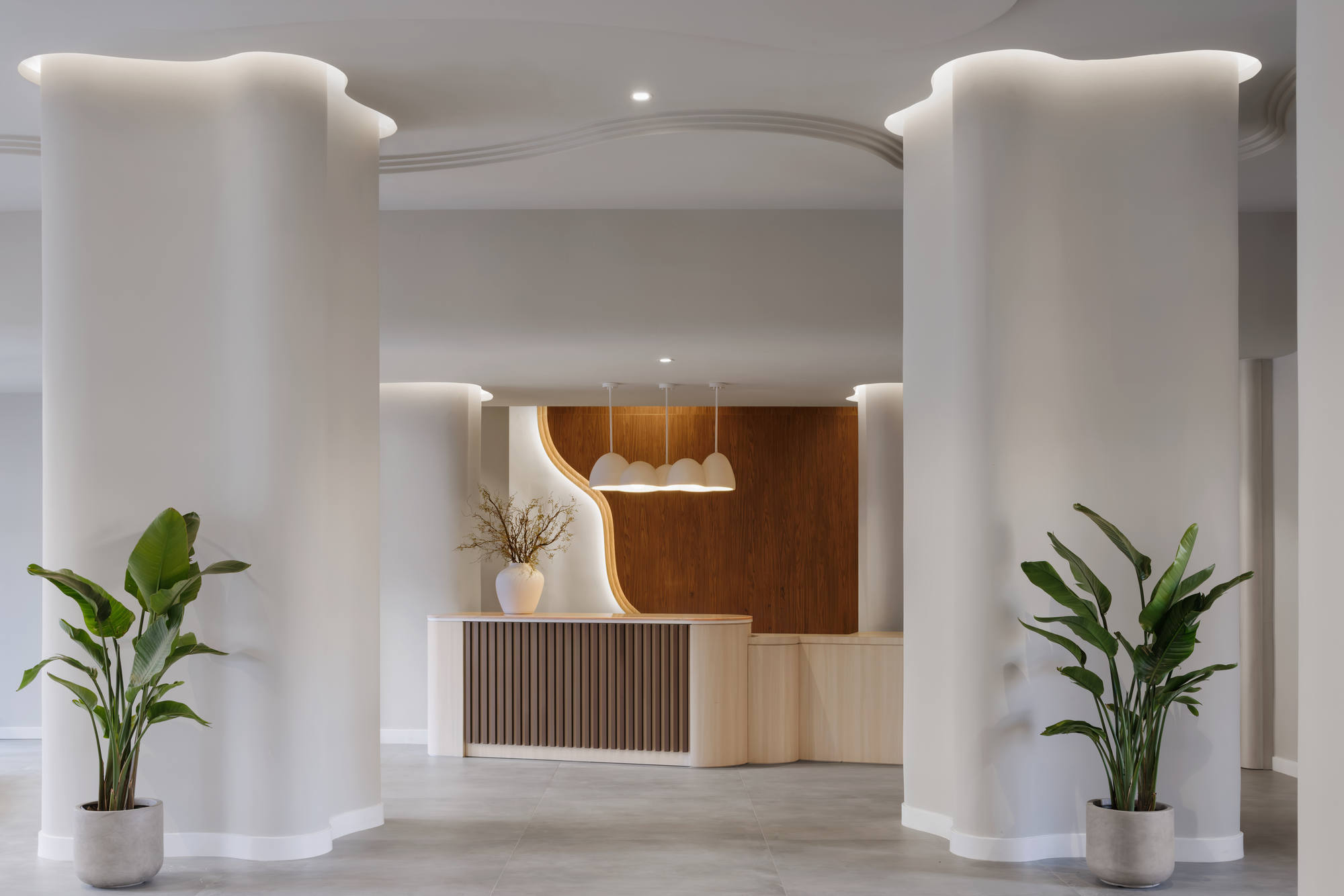 Minimalist building lobby entrance centered on a wood-slat reception desk, framed by massive structural white columns with wave-like curves and embedded cove lighting.