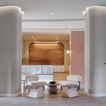 Contemporary lobby sitting area framed by two white columns, highlighting a stylish mailroom alcove with a textured, golden-brown accent wall and minimalist seating arrangements.