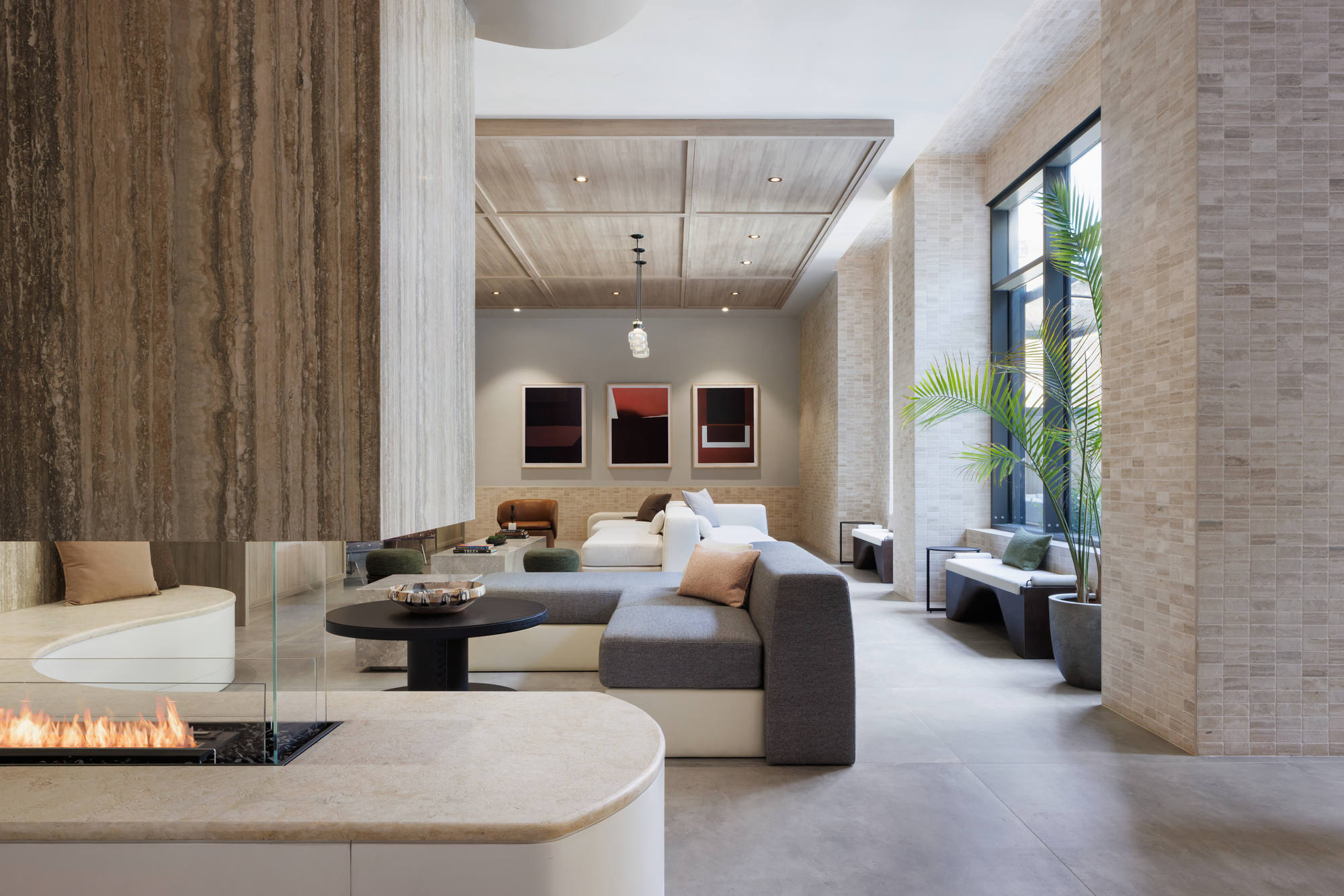 Sophisticated fireplace lounge showcasing a modern glass-enclosed fireplace, plush sectional sofas, textured tile columns, and large windows that let in ample natural light.