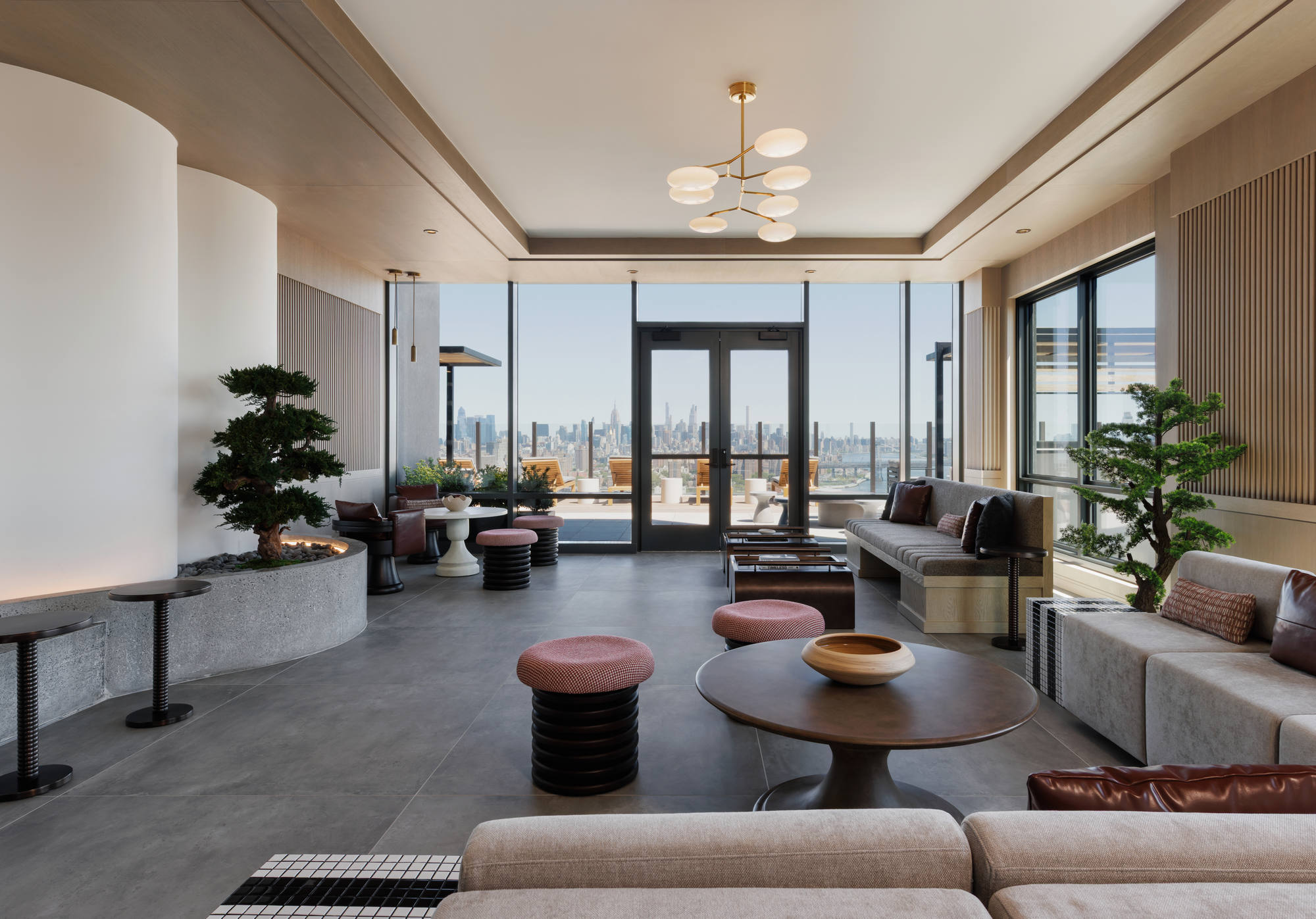 Bright indoor sky lounge with floor-to-ceiling glass windows and doors opening to an outdoor deck, furnished with contemporary low-profile sofas, round tables, and potted bonsai trees.