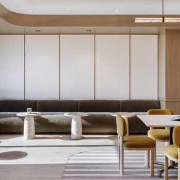 Sleek, minimalist co-working suite featuring velvet ochre chairs around a white marble table, a long low-profile olive green banquette against the back wall, and soft overhead linear lighting.