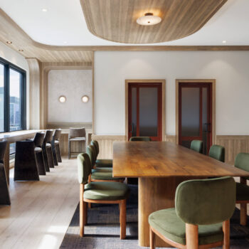 Sophisticated co-working room featuring a large wooden boardroom table with green upholstered chairs, a side counter with tall bar stools beneath a window, and wood-paneled walls and ceiling accents.