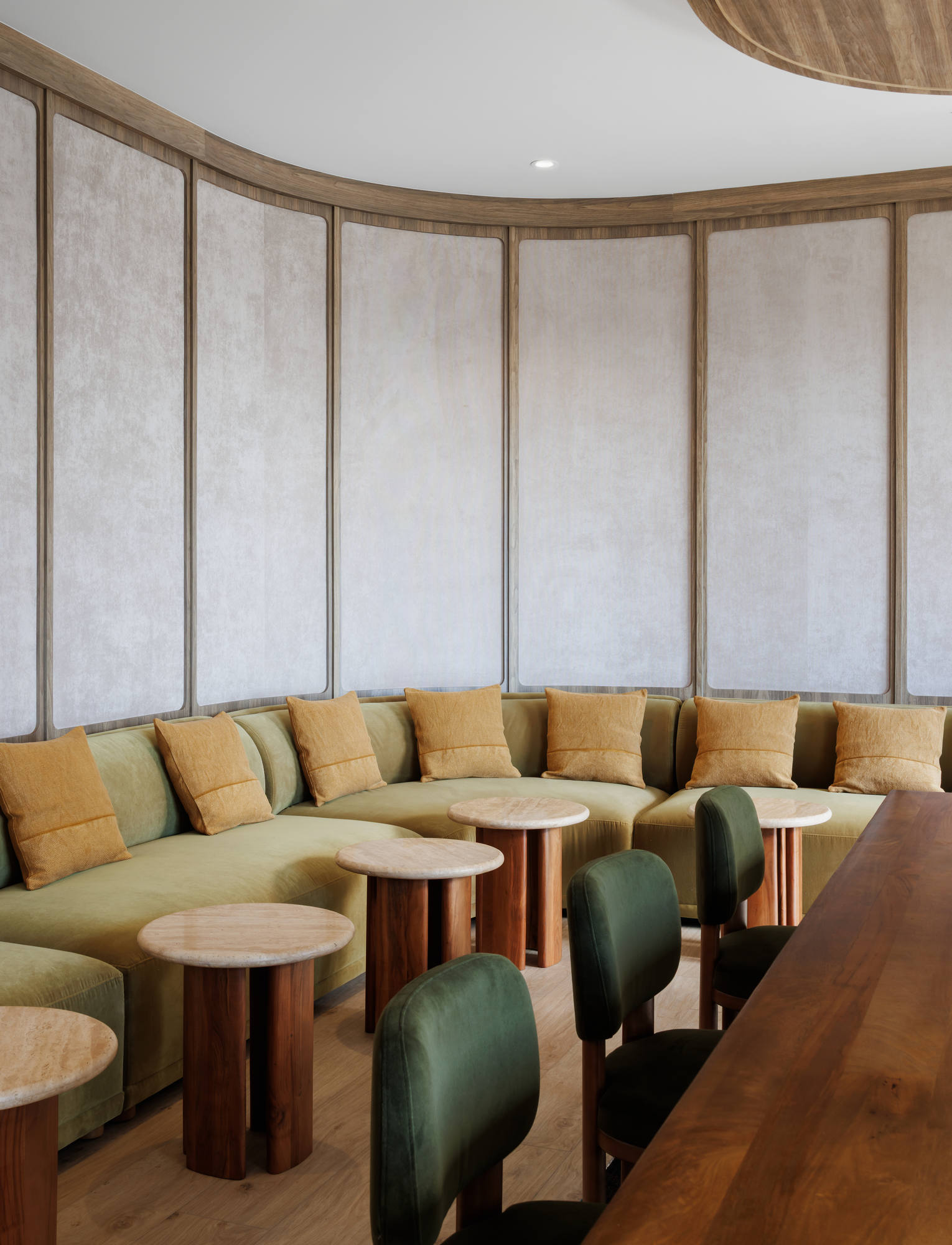 Close-up of a semi-circular booth lounge featuring a sage green velvet banquette accented with mustard yellow pillows and accompanied by small, round stone-topped pedestal tables.