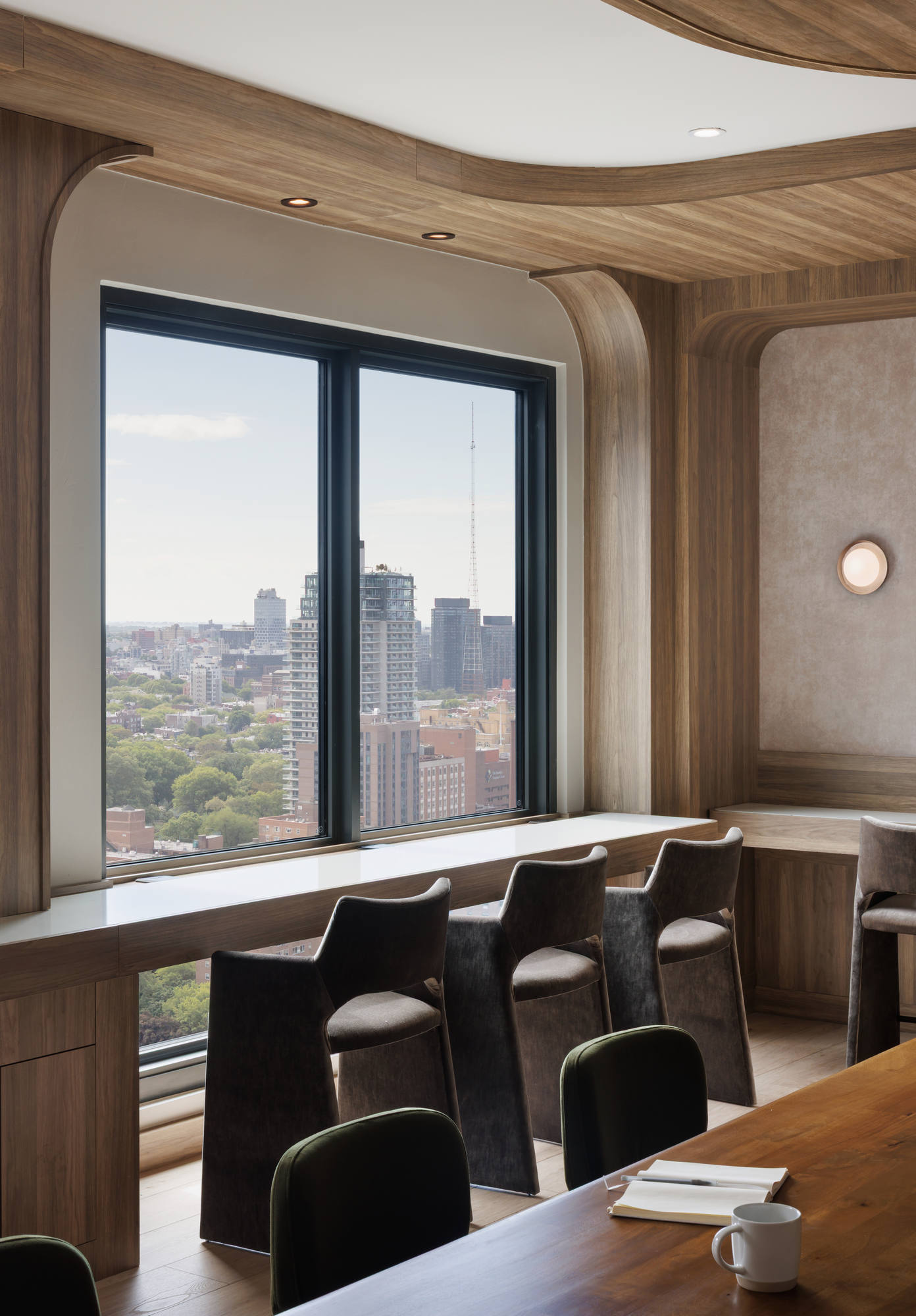 Sleek window-side workspace inside lounge, showing a row of charcoal gray barstools lined up along a built-in wooden counter that overlooks a sweeping city view.