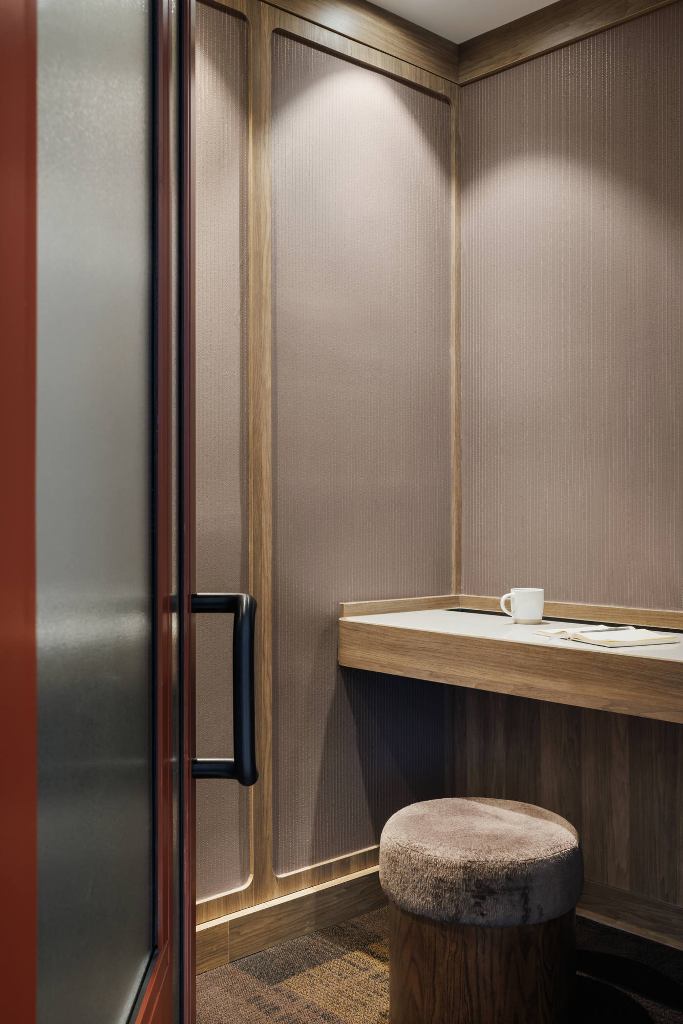 Interior view of a private, wood-paneled are, furnished with a built-in desk, a comfortable plush stool, and textured metallic wallpaper.