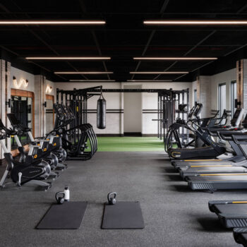 Modern, spacious building gym equipped with a long row of treadmills, stationary exercise bikes, stretching mats with kettlebells, a heavy punching bag, and a green turf functional training area.