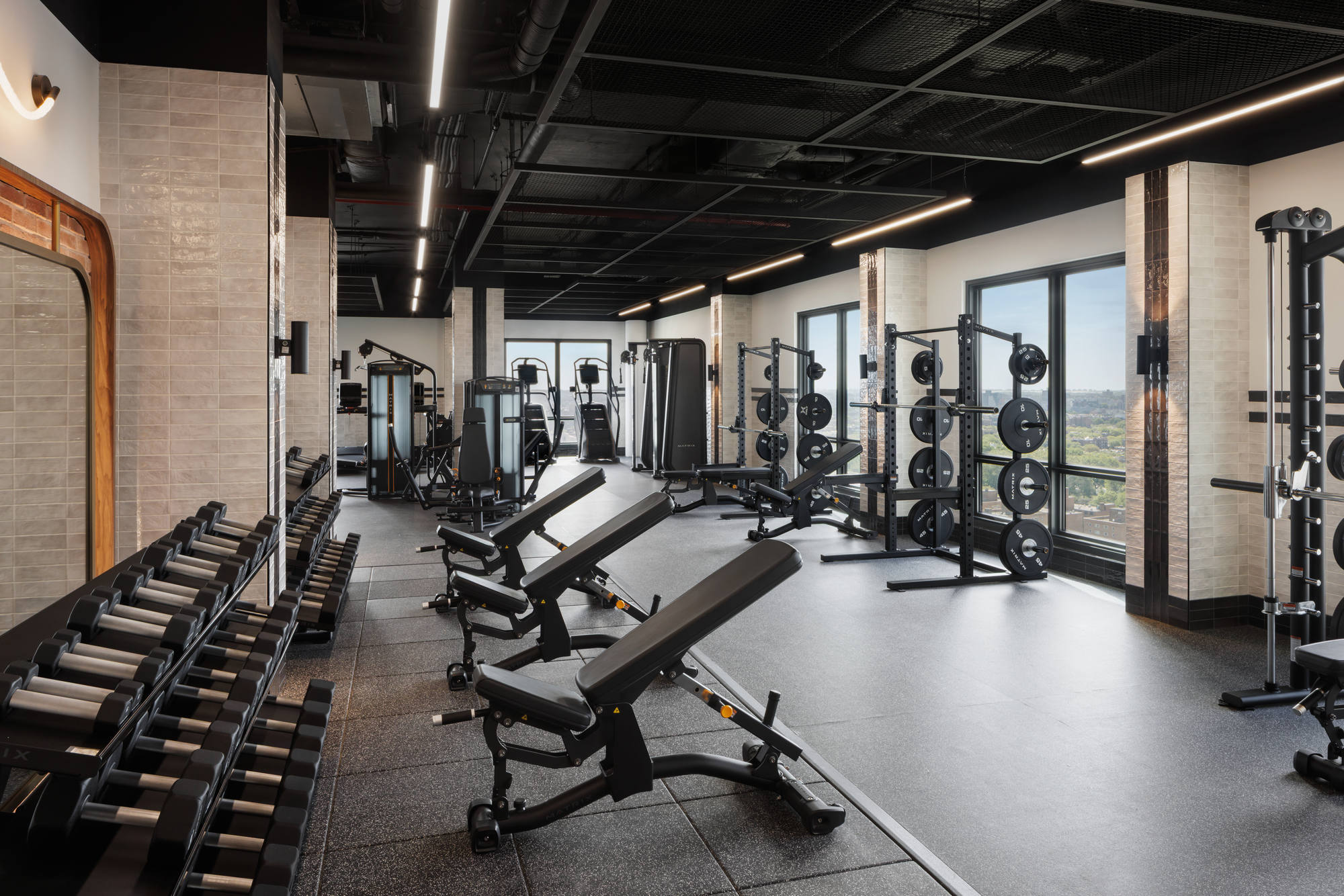Bright, high-end weight room featuring multi-tier dumbbell racks, adjustable weight benches, squat racks with bumper plates, a cable crossover machine, and large windows offering an elevated outdoor view.