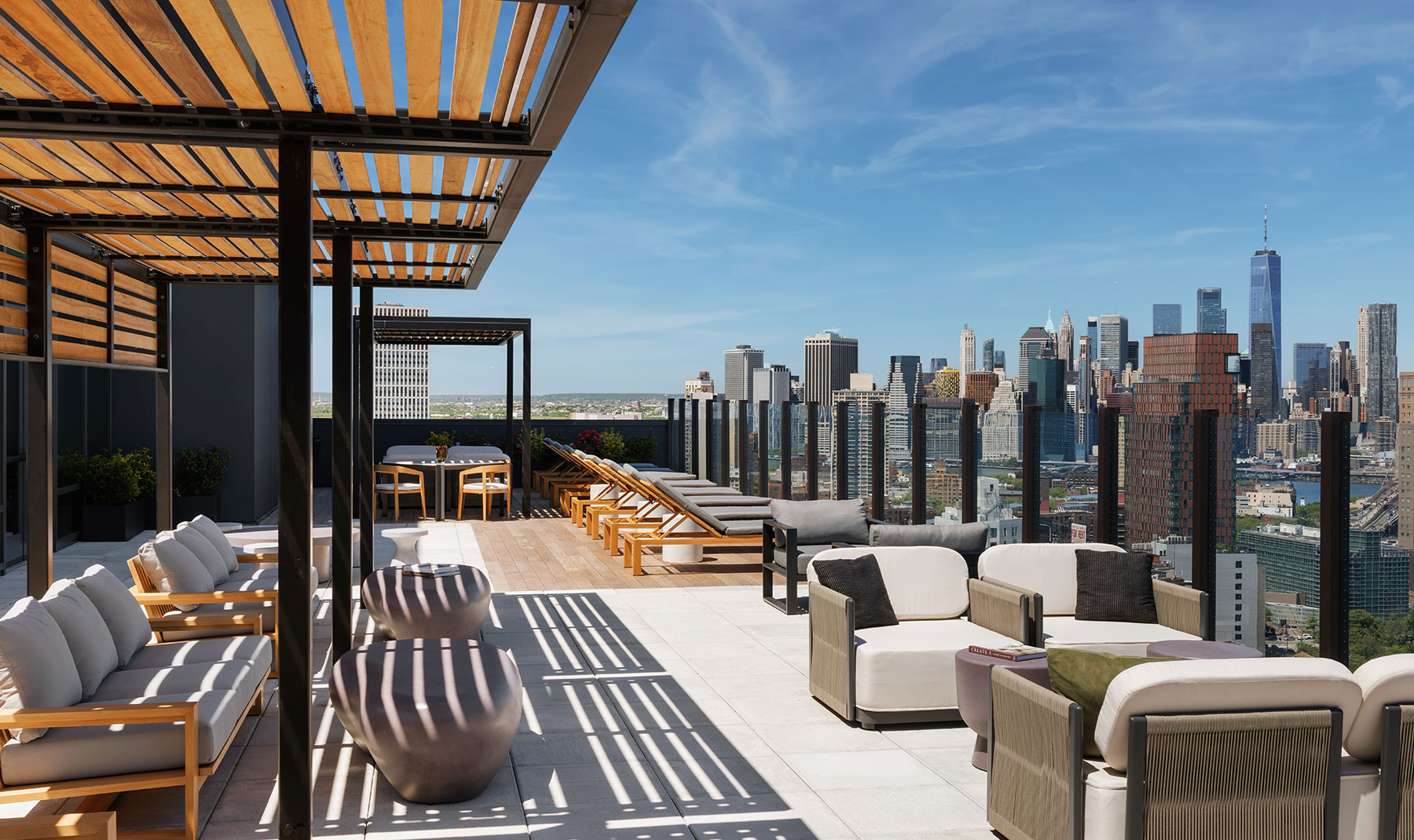 Spacious rooftop amenity deck with modern lounge chairs, sofas, and wooden pergolas, overlooking a panoramic view of the downtown city skyline and One World Trade Center.