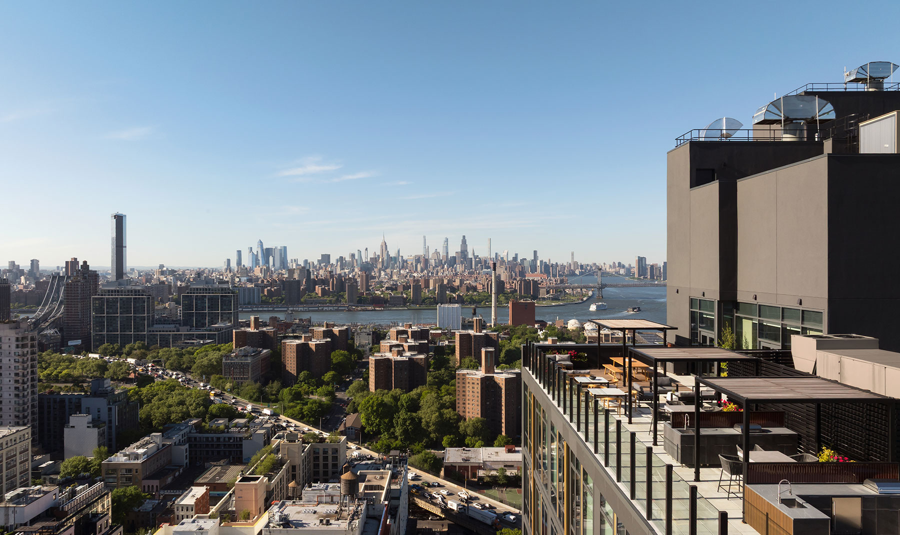 High-angle scenic shot looking past a luxury building's multi-level rooftop terrace down toward a bustling urban landscape, tree-filled parks, and a distant city skyline under a clear sky.
