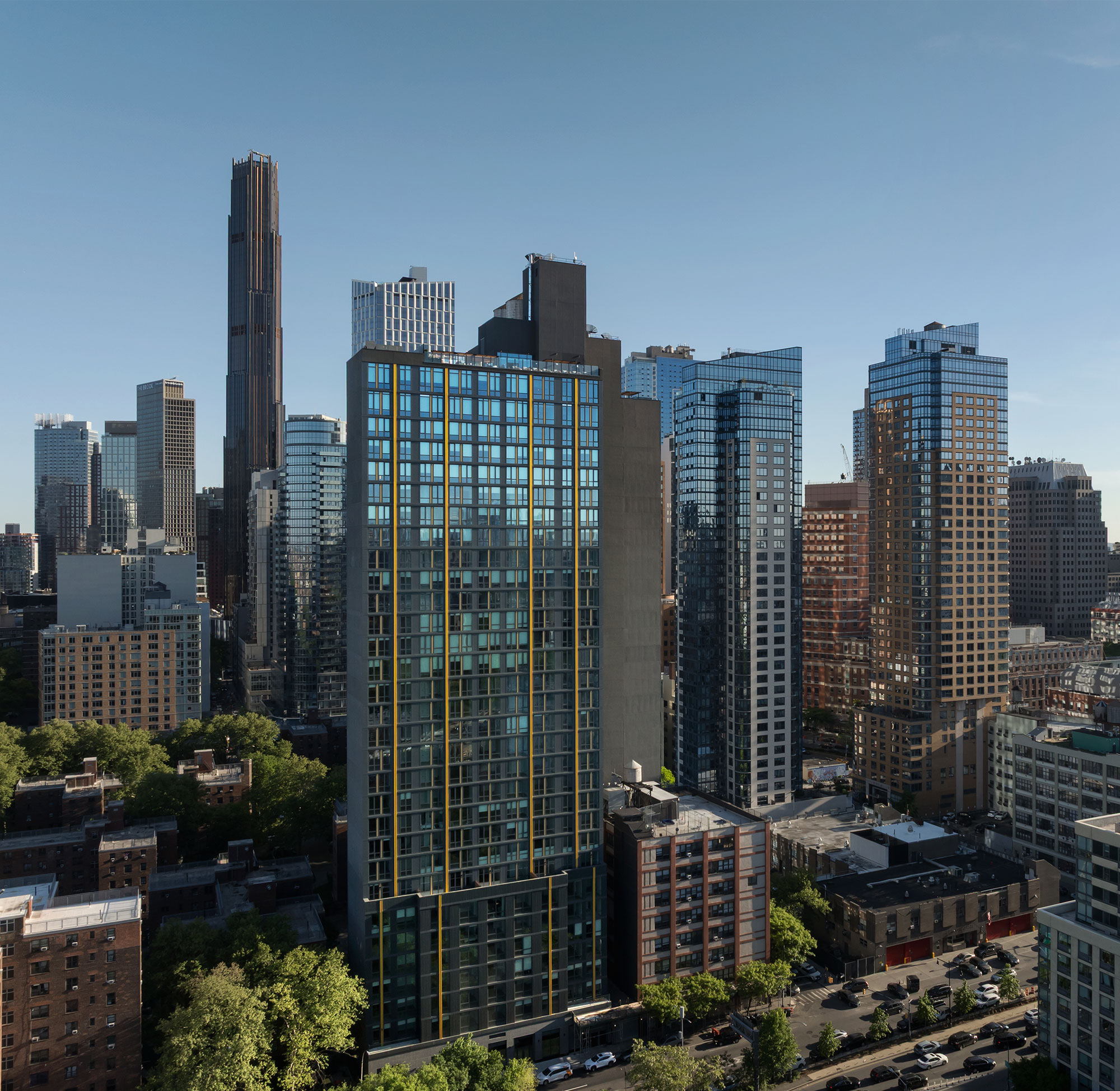 Aerial shot focusing on the dark high-rise rental tower with its prominent vertical gold stripes, nestled amongst neighboring skyscrapers and lush green trees under a clear blue sky.