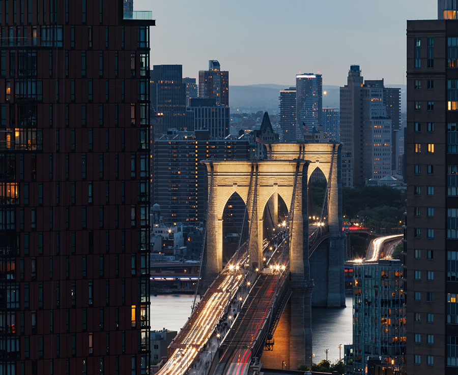 Telephoto architectural shot capturing the historic stone arches of the Brooklyn Bridge at dusk, illuminated by bright streams of vehicle headlights and framed by modern skyscrapers.